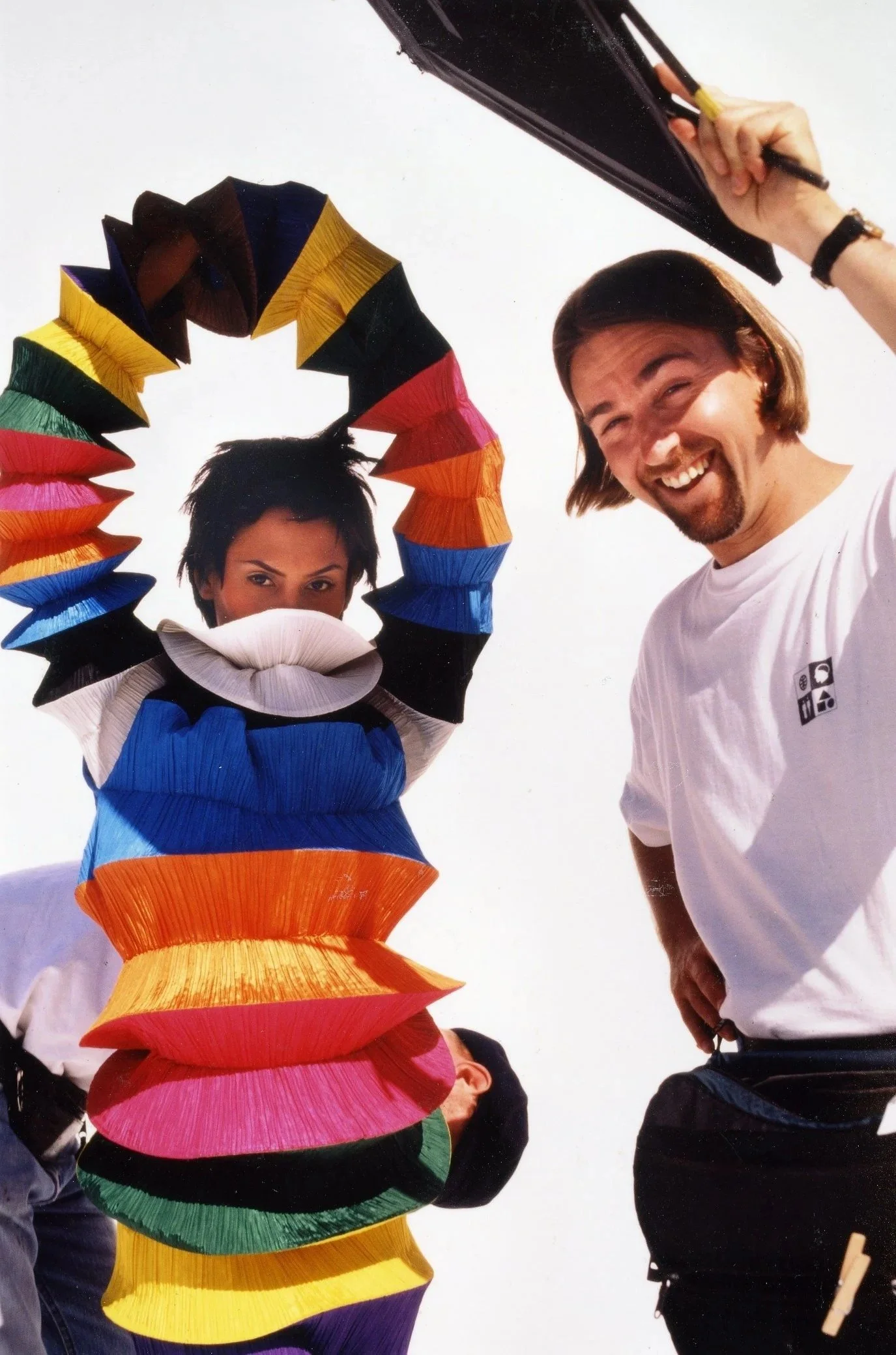 People standing outdoors, one person with a large, colorful, spiral sculpture made of stacked paper plates, and another person smiling and holding a black object.