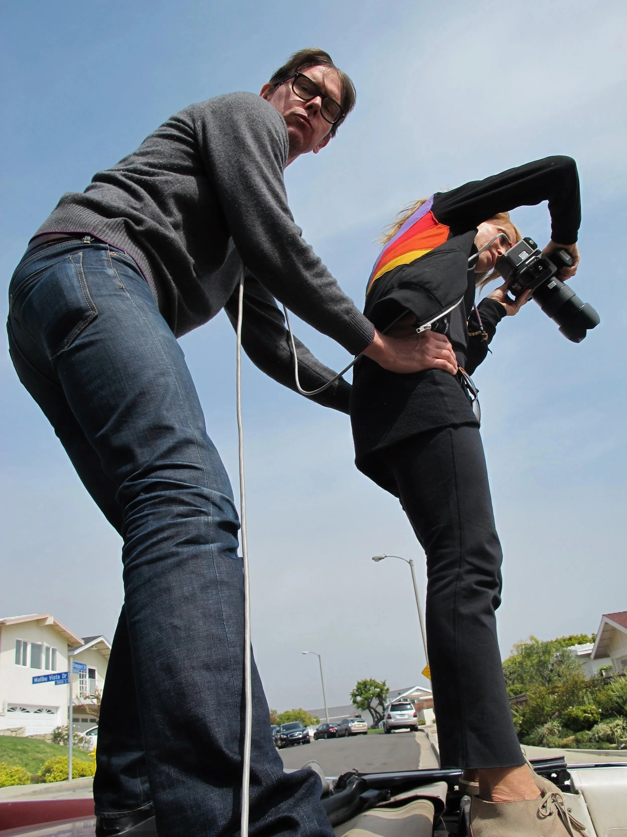 Two people appear to be standing on a vehicle with houses and parked cars in the background, with one person leaning over and the other looking down.