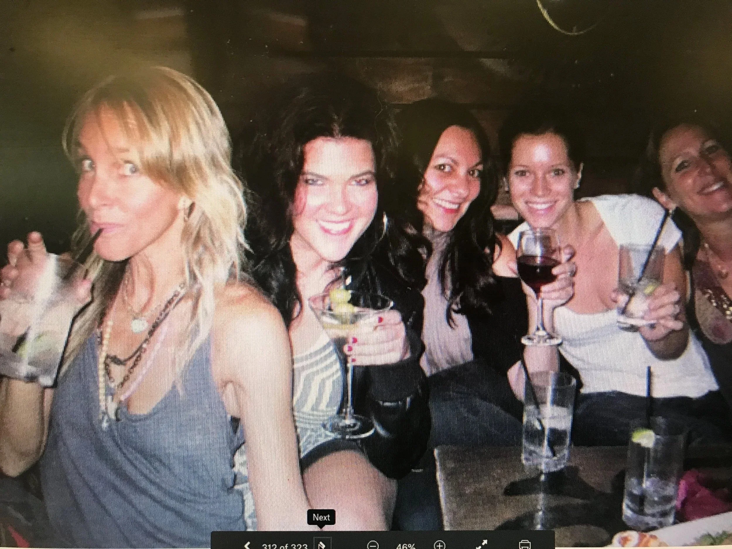 A group of five women at a social gathering, smiling and holding drinks, with a dark background and a patterned tablecloth in front of them.