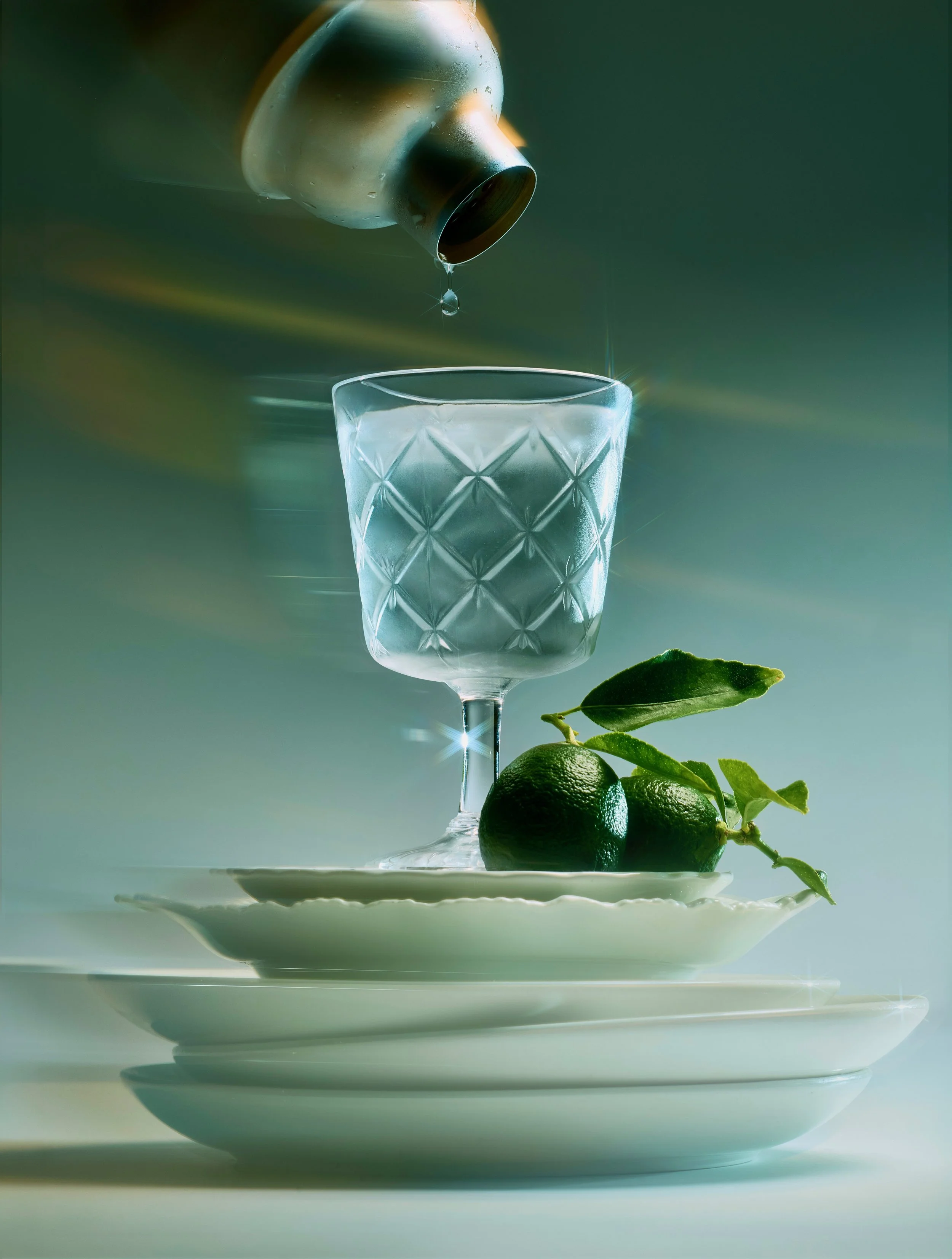 A clear wine glass with a textured diamond pattern, on a white plate, with a lemon and green leaves beside it. A bottle tips pouring a dark liquid into the glass, with a droplet falling and a soft green background.