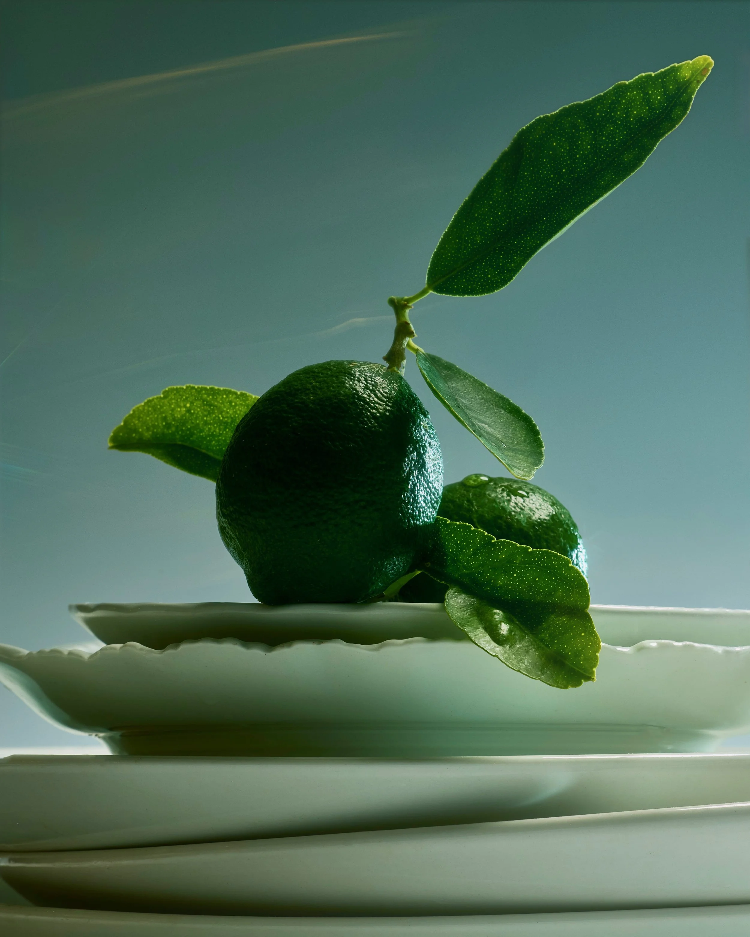 Close-up of a stack of three green limes on a white plate with a grey background, a lime leaf attached at the top.