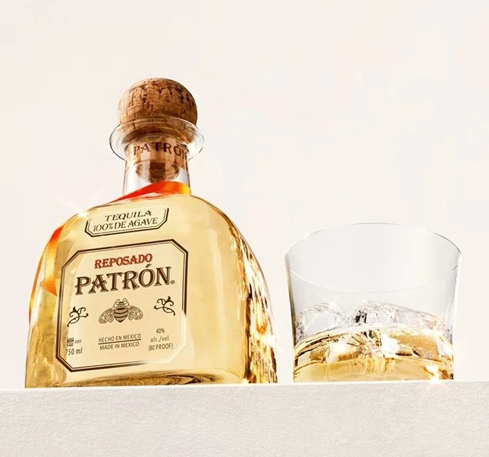 A bottle of Patrón Reposado tequila with a glass of tequila and ice beside it on a white surface.