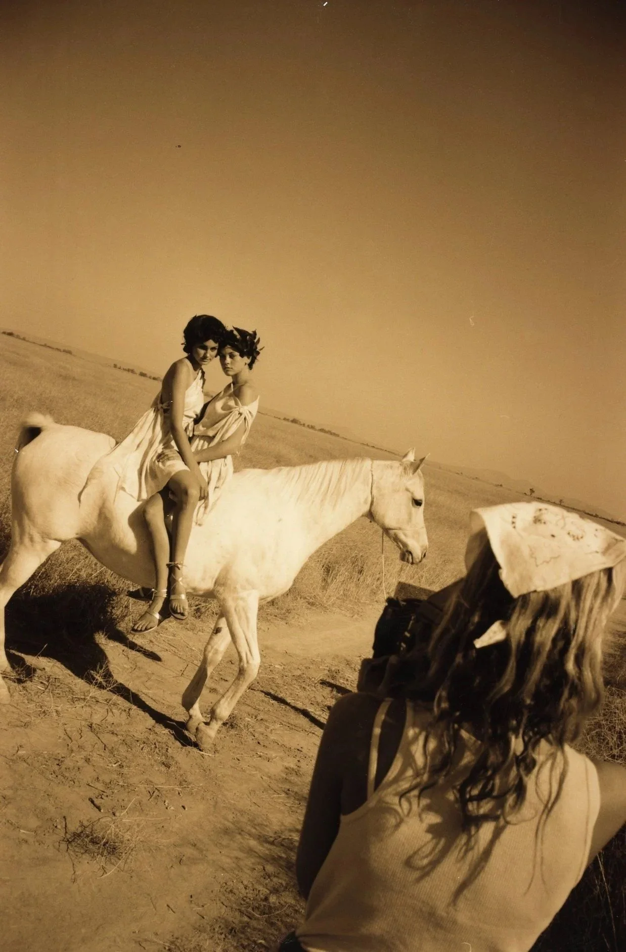 Two women sitting on a white horse in a desert landscape at dusk, with a person in the foreground wearing a hat and long hair.