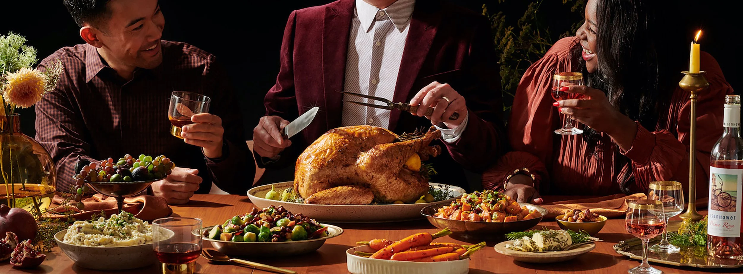 Three people enjoying a Thanksgiving dinner with a roasted turkey, side dishes, and drinks at the table. One person is carving the turkey while the others are holding glasses and smiling.