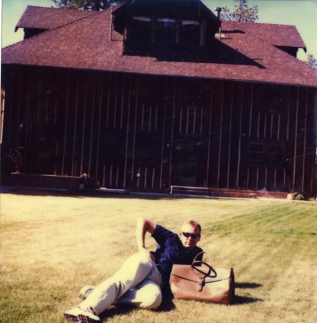 A man with sunglasses lying on the grass in front of a large wooden house with a red shingle roof and a small upper balcony. The man has a tan bag beside him.
