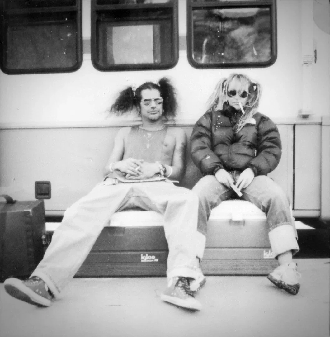 Two young women sitting on a bench outside a vehicle, with one wearing sunglasses and a puffy jacket and the other with glasses and messy hair, both appearing relaxed.