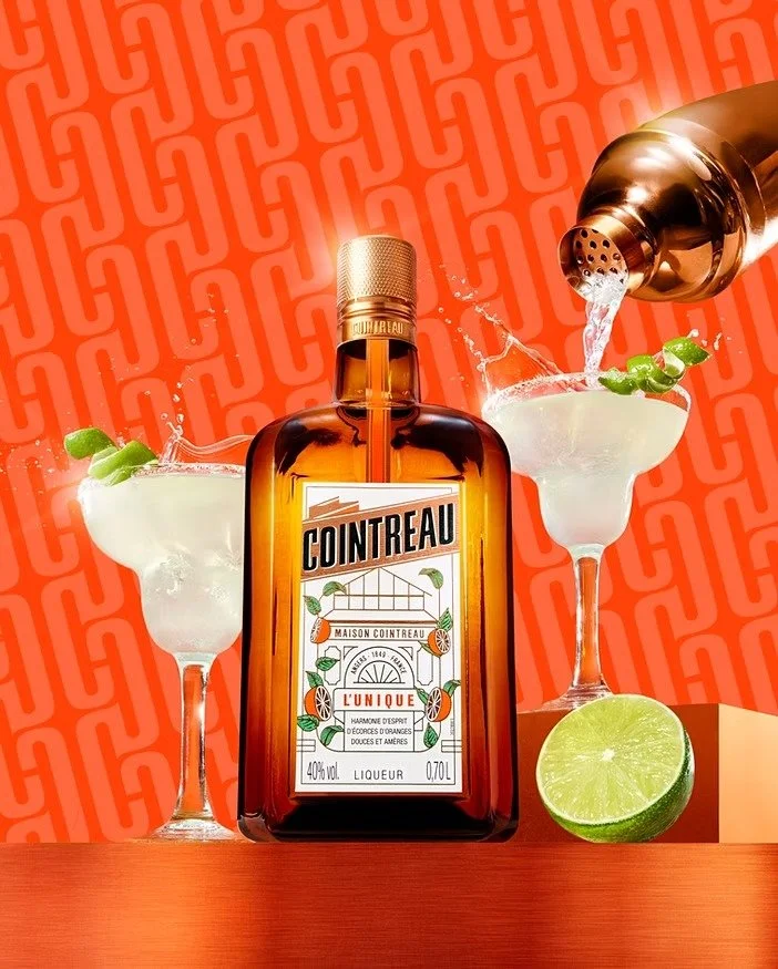 A bottle of Cointreau liqueur with two cocktail glasses, a lime, and a shaker pouring liquid into one glass, against a patterned orange background.