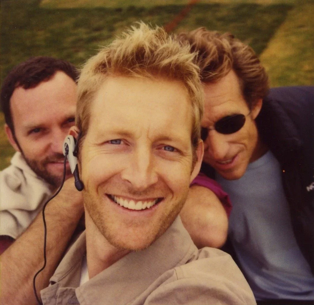 Three men smiling for a selfie outdoors, one wearing a headset, another with sunglasses, on a grassy background.