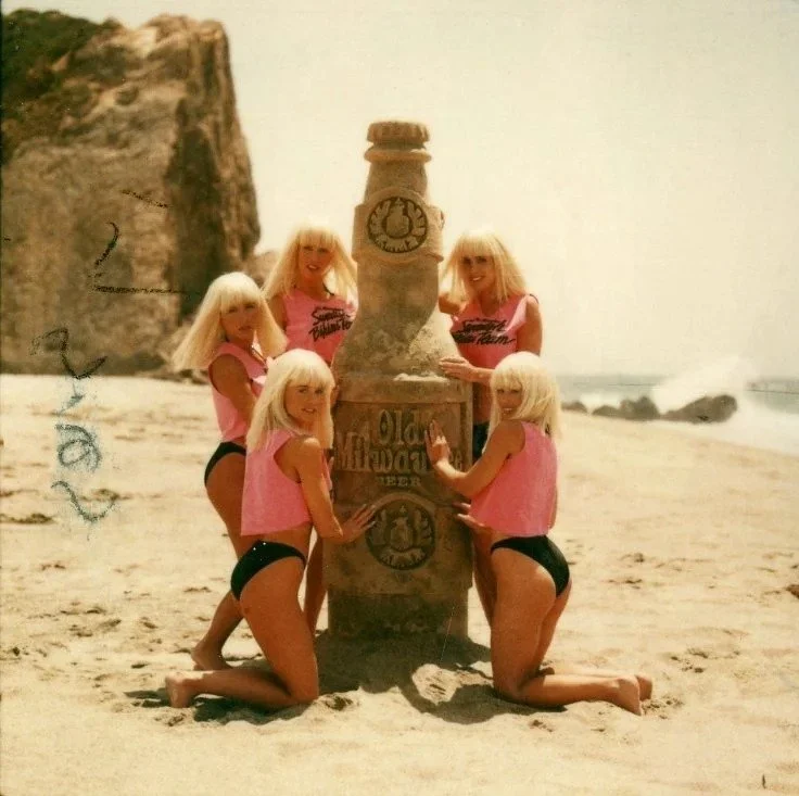 Five women with blonde hair and pink Tank Tops and black high-waist bottoms posing on a beach with a large Old Worster beer bottle statue.