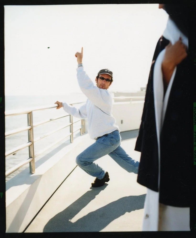 A man with dark curly hair, sunglasses, and a baseball cap is striking a playful pose on a pier, holding onto the railing with one hand and raising his other arm with his index finger pointing upward.