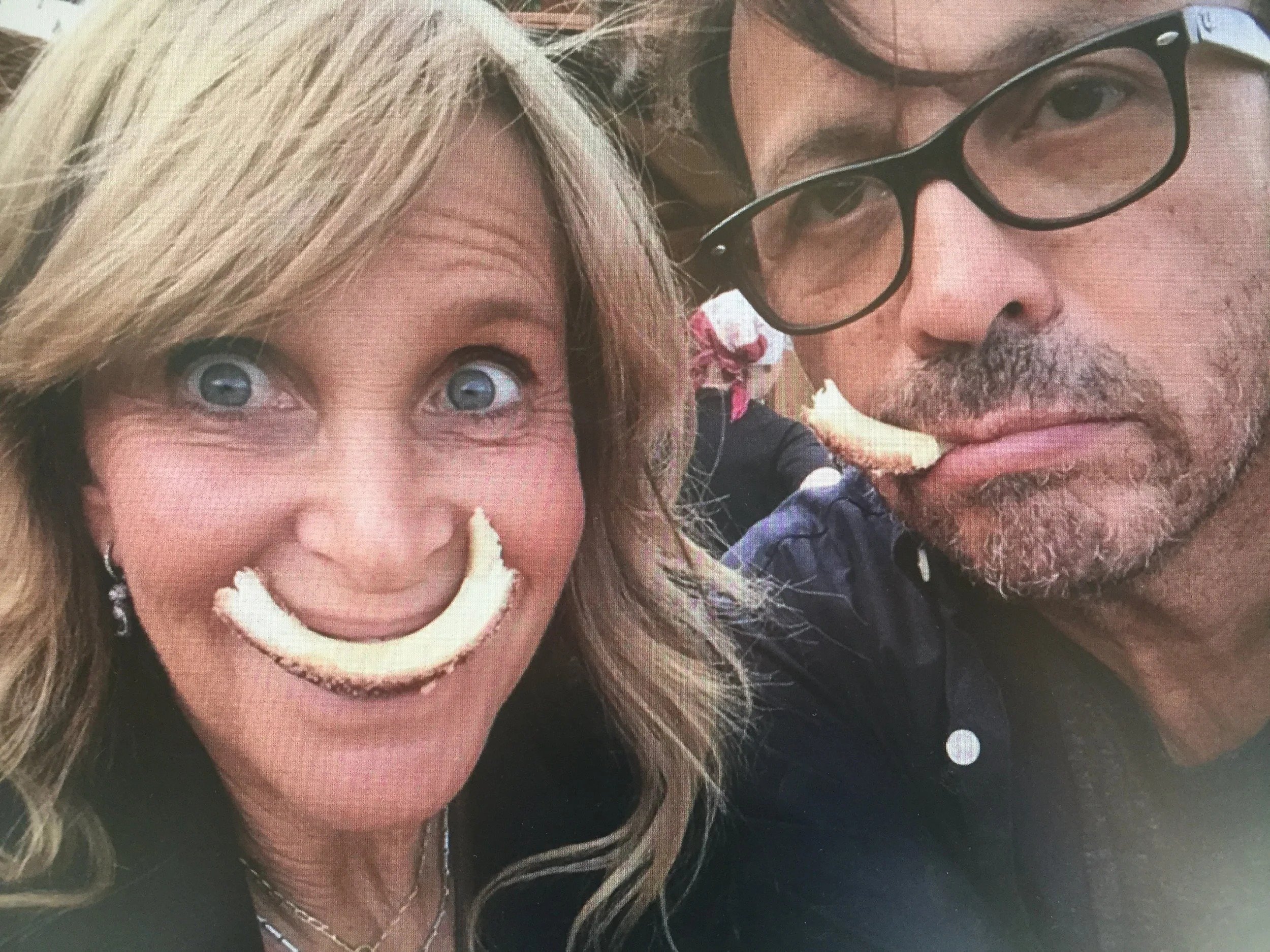 Close-up of a smiling woman with blonde hair and blue eyes, holding a slice of cake near her mouth, and a man with glasses and a beard, looking at the camera, both appearing to enjoy a celebration.