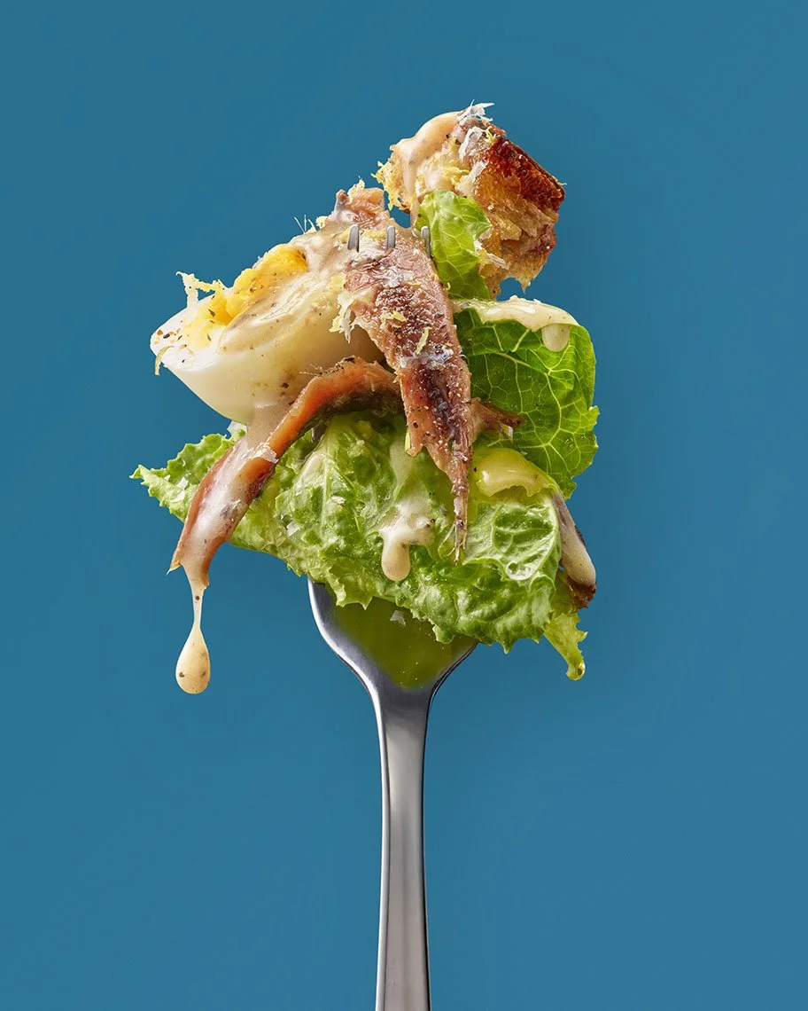 A fork with a bite-sized portion of Caesar salad including lettuce, bacon, boiled egg, and creamy dressing, set against a plain blue background.