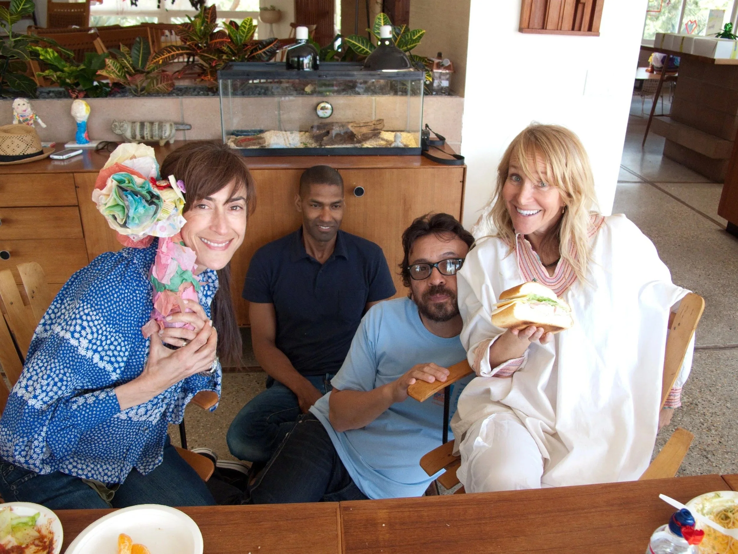 A group of five people sitting around a wooden table in a restaurant, smiling at the camera. Two women are holding food and wearing colorful outfits, while three men are sitting beside them. The table has plates of food, and there is a small animal h