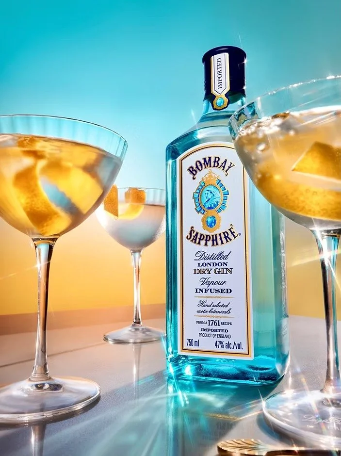A bottle of Bombay Sapphire gin surrounded by three cocktail glasses filled with gin and lemon slices. The bottle is blue with a white label and gold accents, set against a gradient background from blue to yellow.