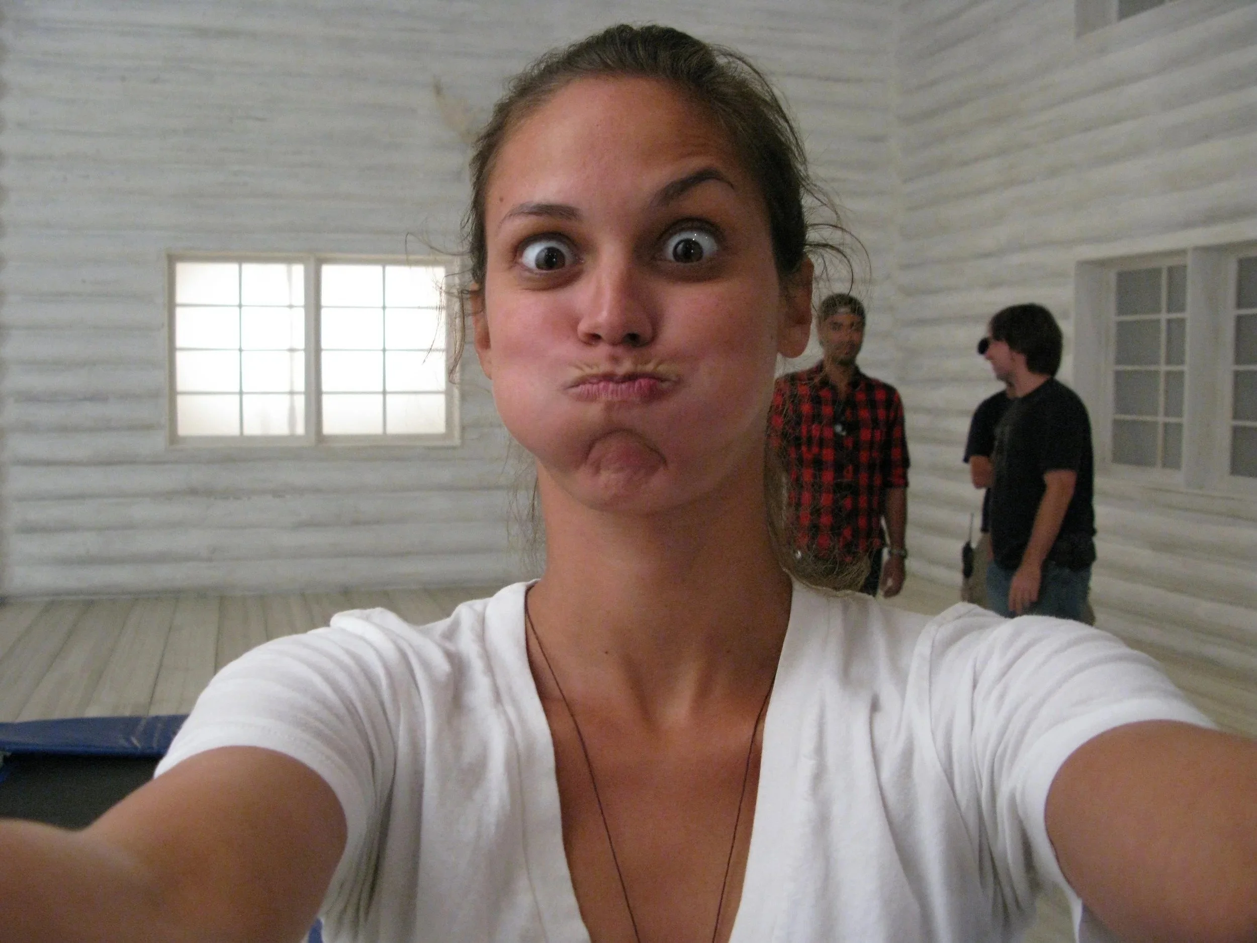 A woman taking a selfie with two men in the background inside a room with white wooden walls and large windows.