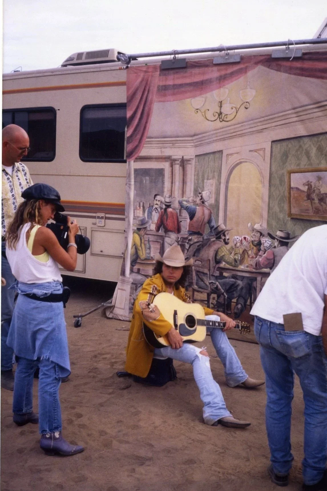 A man in a yellow shirt and a hat is sitting on a stool playing an acoustic guitar at an outdoor event. Several people are around him, with a person in a white tank top and ripped jeans taking a picture or video, and another person with a camera. Beh