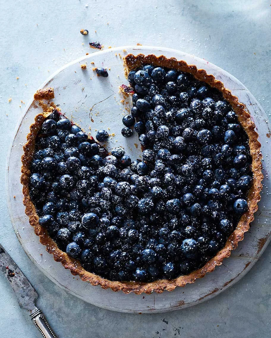A blueberry tart with a portion missing, on a round glass plate, with scattered blueberries and crumbs around it.