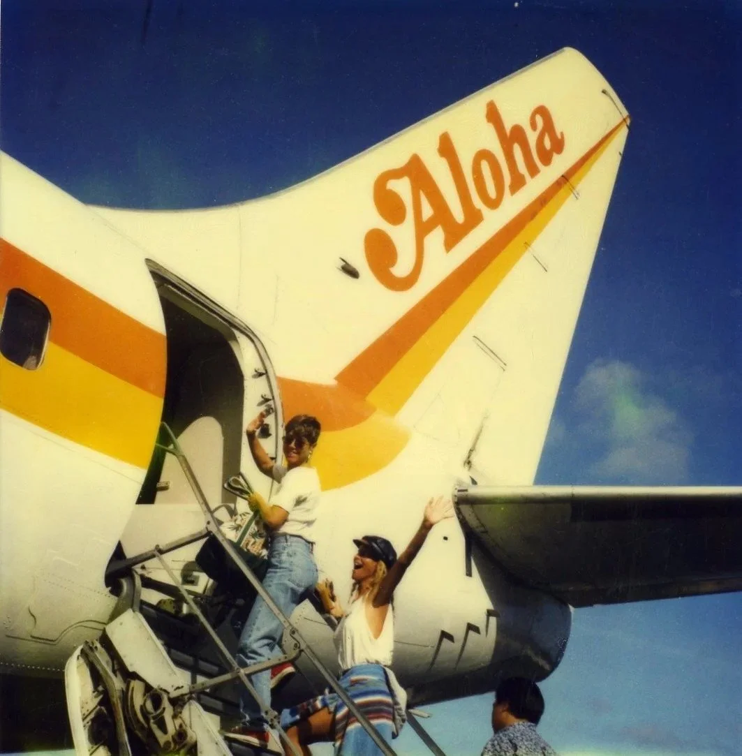 People boarding a Hawaiian Airlines airplane on the tarmac, with a woman waving and smiling.