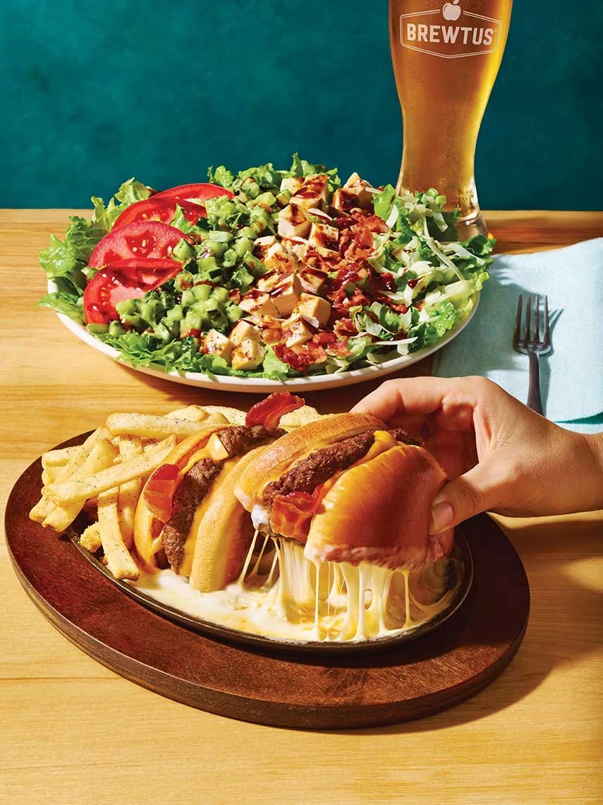 A cheeseburger with fries on a sizzling platter, a large salad with tomatoes, cucumbers, shredded cheese, and chopped meats, a tall glass of beer, and a fork on a table.