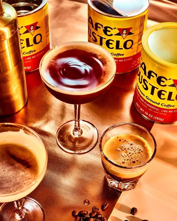 A variety of coffee drinks and coffee cans on a table, including a martini glass with a dark liquid, small glass with crema, and canned ground coffee on a shiny surface.