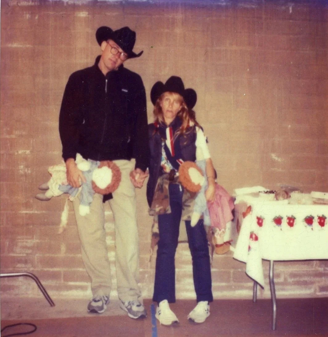 A man and a woman dressed in cowboy hats and casual clothing standing in front of a brick wall, holding children and their hats. A table with a holiday-themed tablecloth is visible on the right side of the image.
