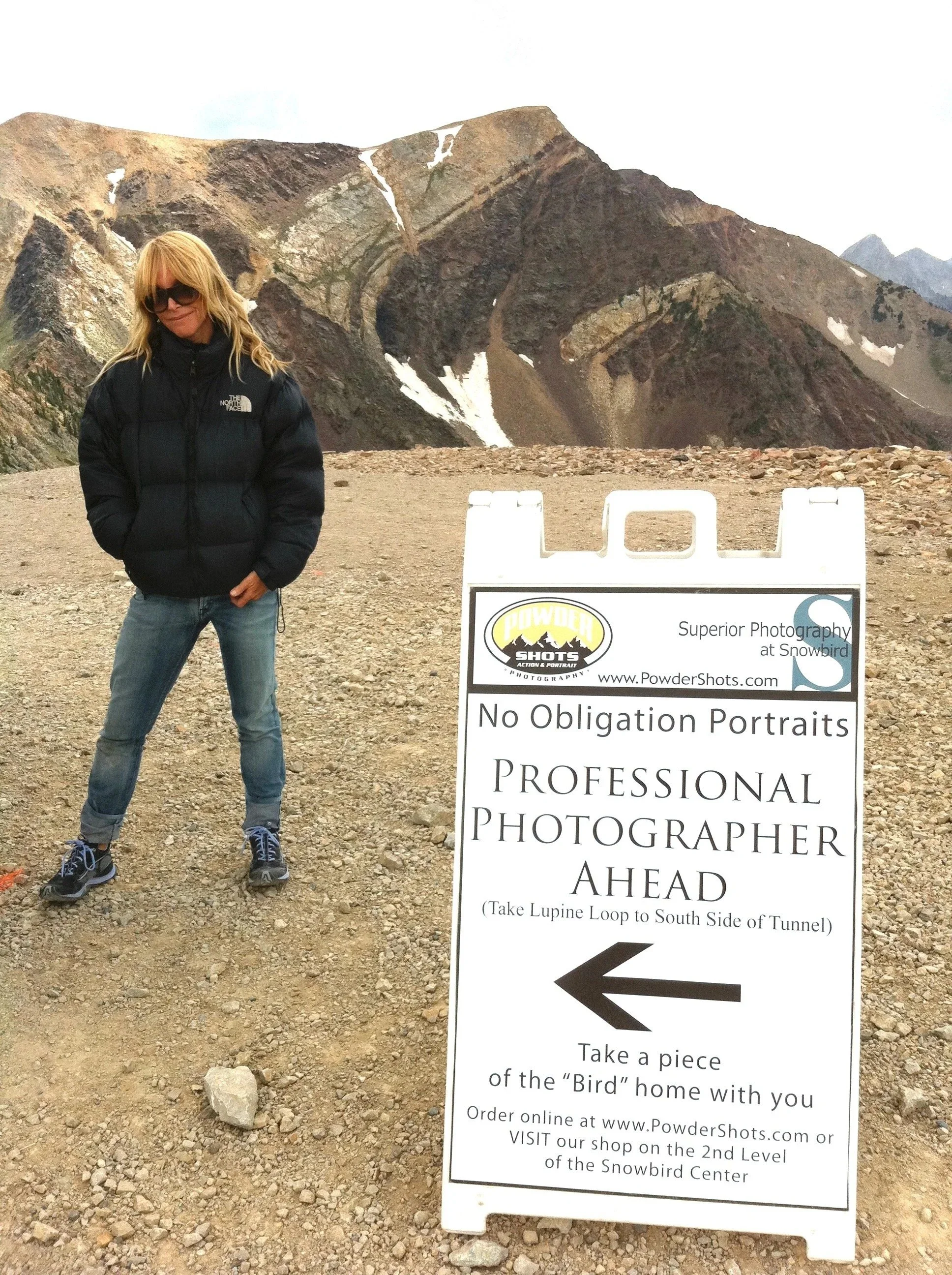 A woman with blonde hair wearing sunglasses and a black North Face puffer jacket standing next to a large sign at Snowbird mountain. The mountain behind her is rocky with patches of snow. The sign indicates an area with no obligation for portraits an