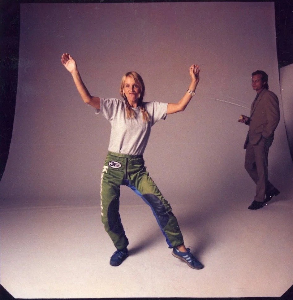 An older woman in a casual outfit, including a gray t-shirt and green racing pants, is posing with her arms raised and slightly bent at a dance or celebration. A man in a gray suit stands in the background, holding an object, possibly a camera or rem