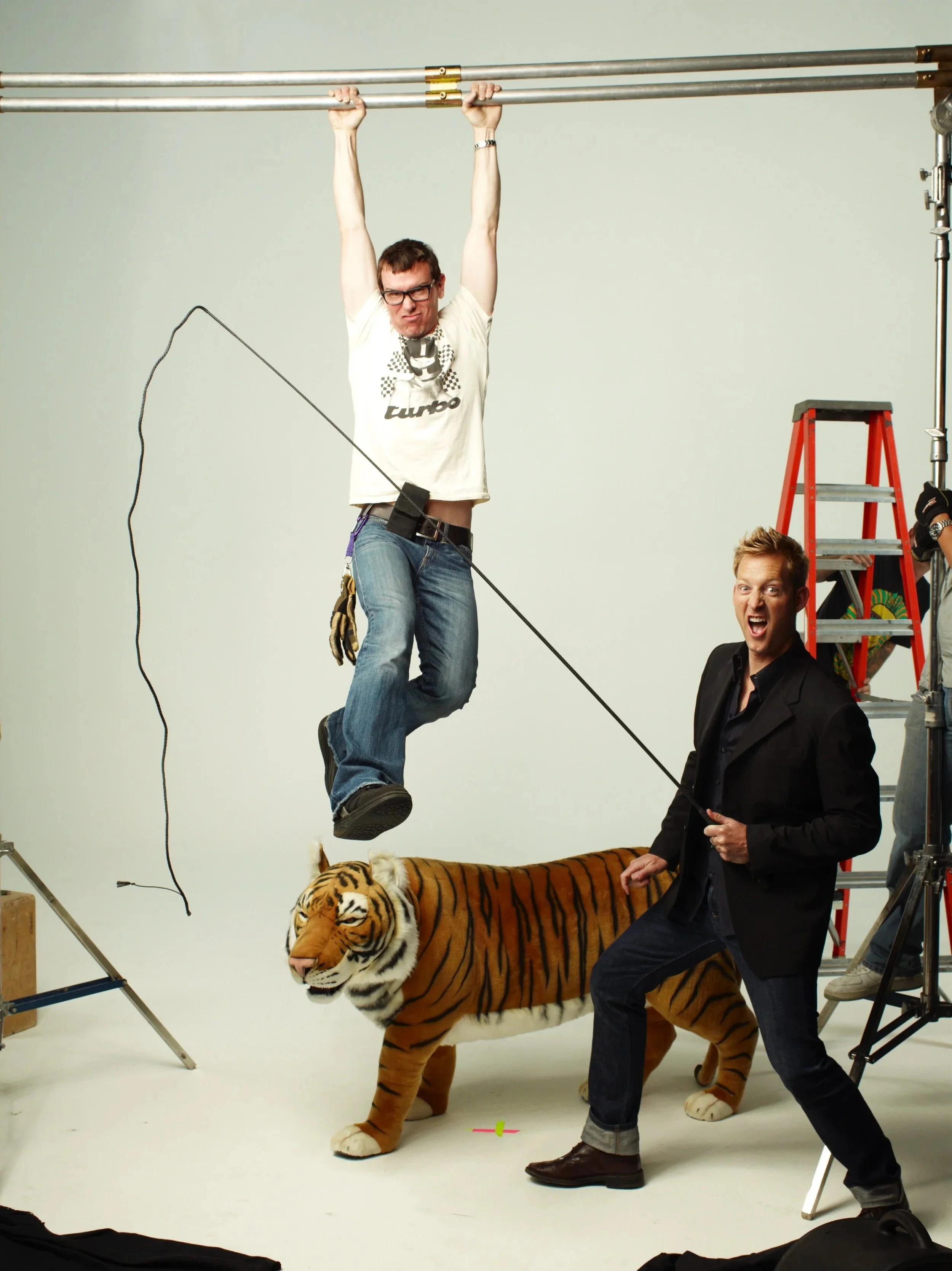 People posing with a tiger prop and a person hanging from a scaffold in a photo studio, with equipment and a ladder visible.
