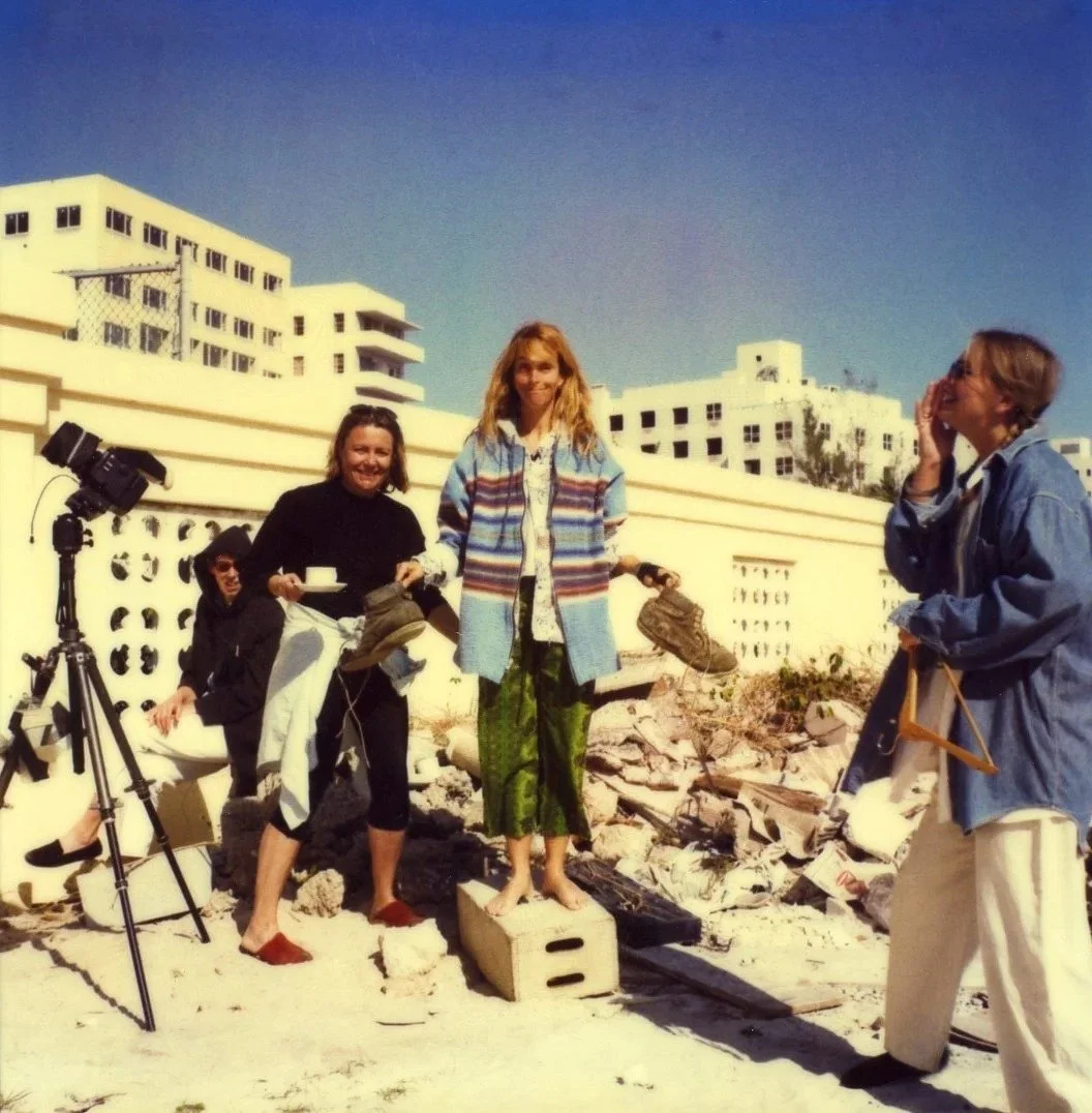 A woman stands on a wooden crate among rubble, with a woman to her right laughing and a woman to her left holding a cup. There are others and a camera on a tripod, with high-rise buildings in the background.