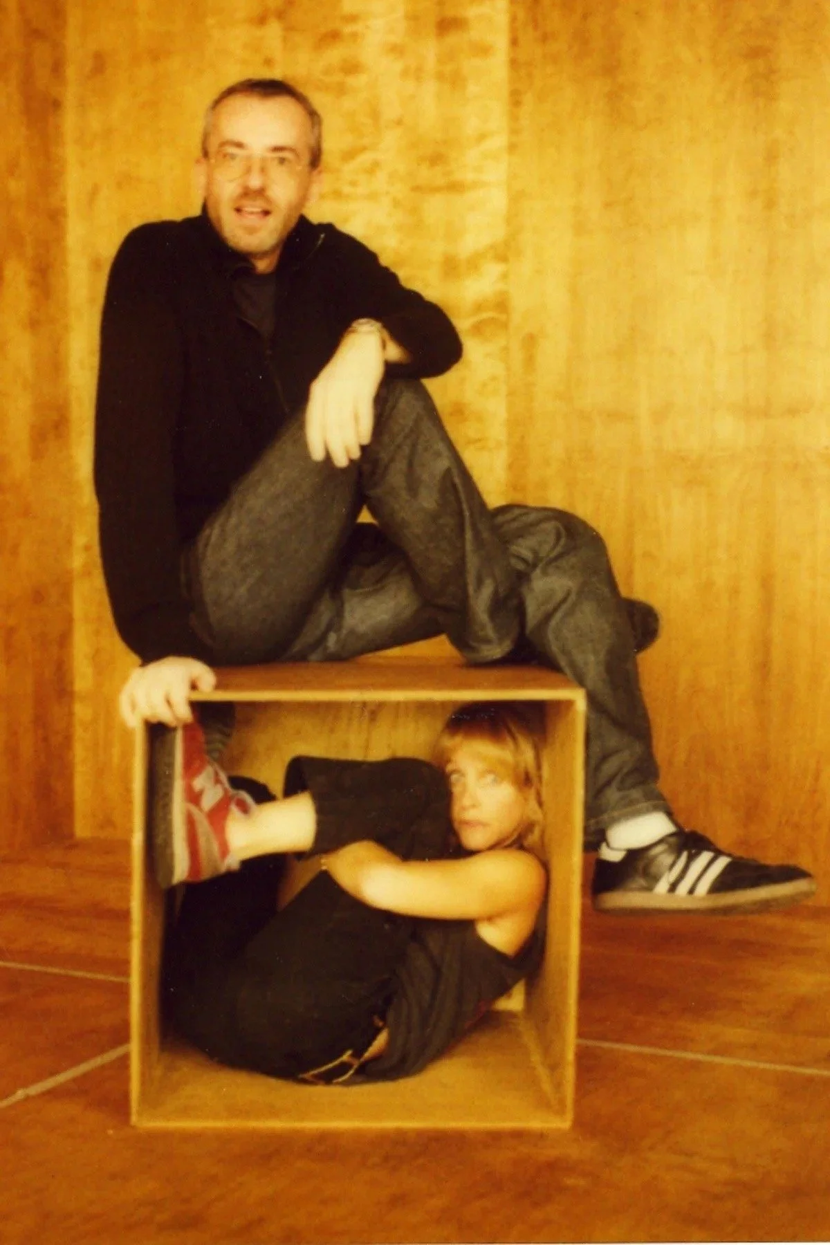 Two children inside a wooden cube, with one sitting on the floor inside the cube and the other on top of it. An adult man sits atop the cube, with one leg on the cube's edge and the other extended. The background is wooden paneling.