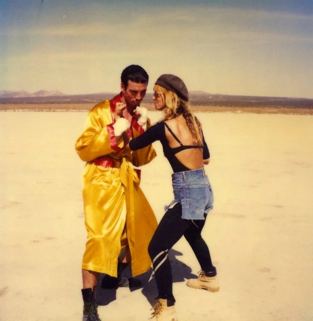A man and a woman appear to be boxing or fighting in a desert landscape, with the man wearing a shiny yellow robe and the woman dressed in a black top, ripped denim shorts, and combat boots.