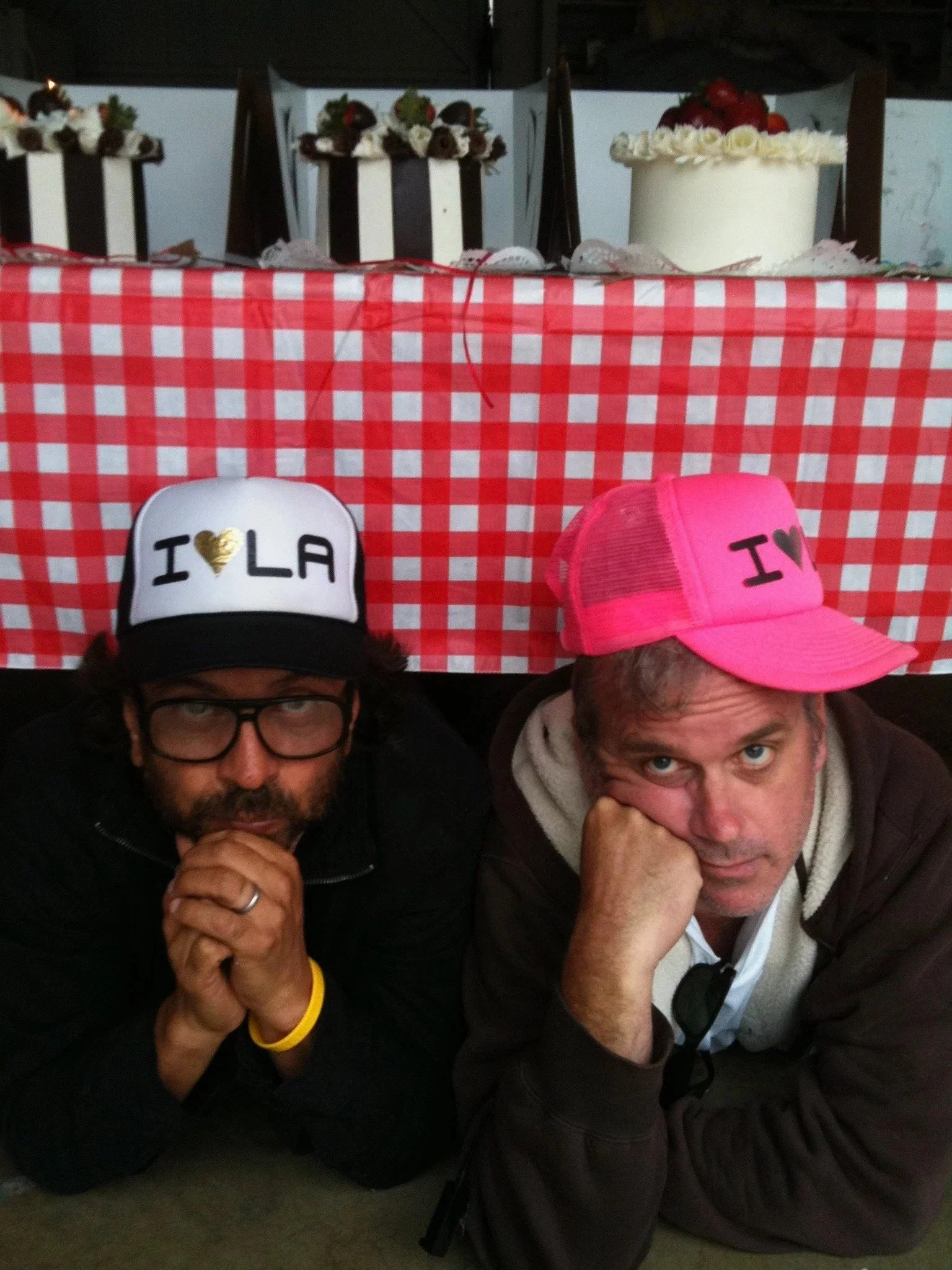 Two men are lying on the ground, leaning on their elbows, under a table with three cakes on top. The man on the left wears a white trucker hat with a gold heart and the words 'I LA', and a black shirt. The man on the right wears a pink trucker hat wi