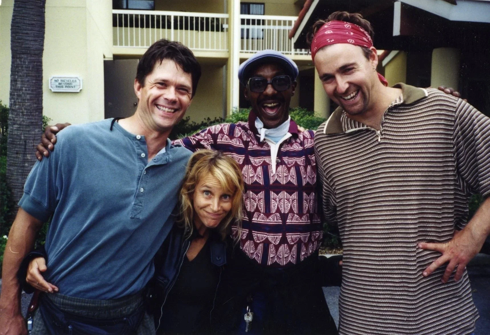 Four people standing close together outside a building, smiling and posing for a photo. One man is wearing a gray shirt, another man in a colorful patterned shirt with glasses, a woman has long blonde hair, and another man is wearing a striped shirt 