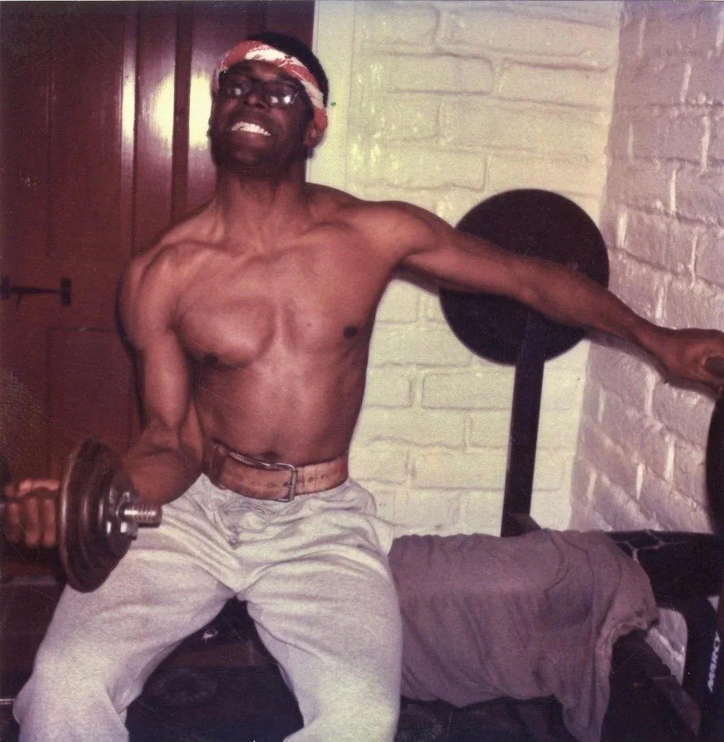 A shirtless man with glasses and a bandana, smiling while lifting a dumbbell. He is sitting on a bench in a room with a white brick wall and wooden door.