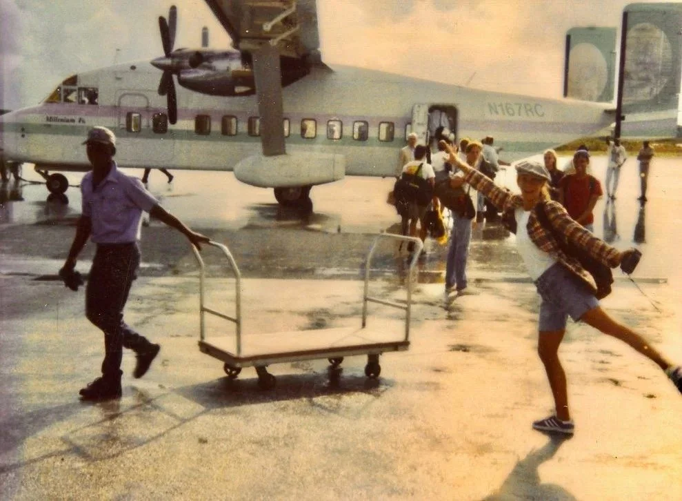 People arriving at an airport with a turboprop airplane in the background, some pulling luggage carts, and a girl playfully posing with outstretched arms in the foreground.