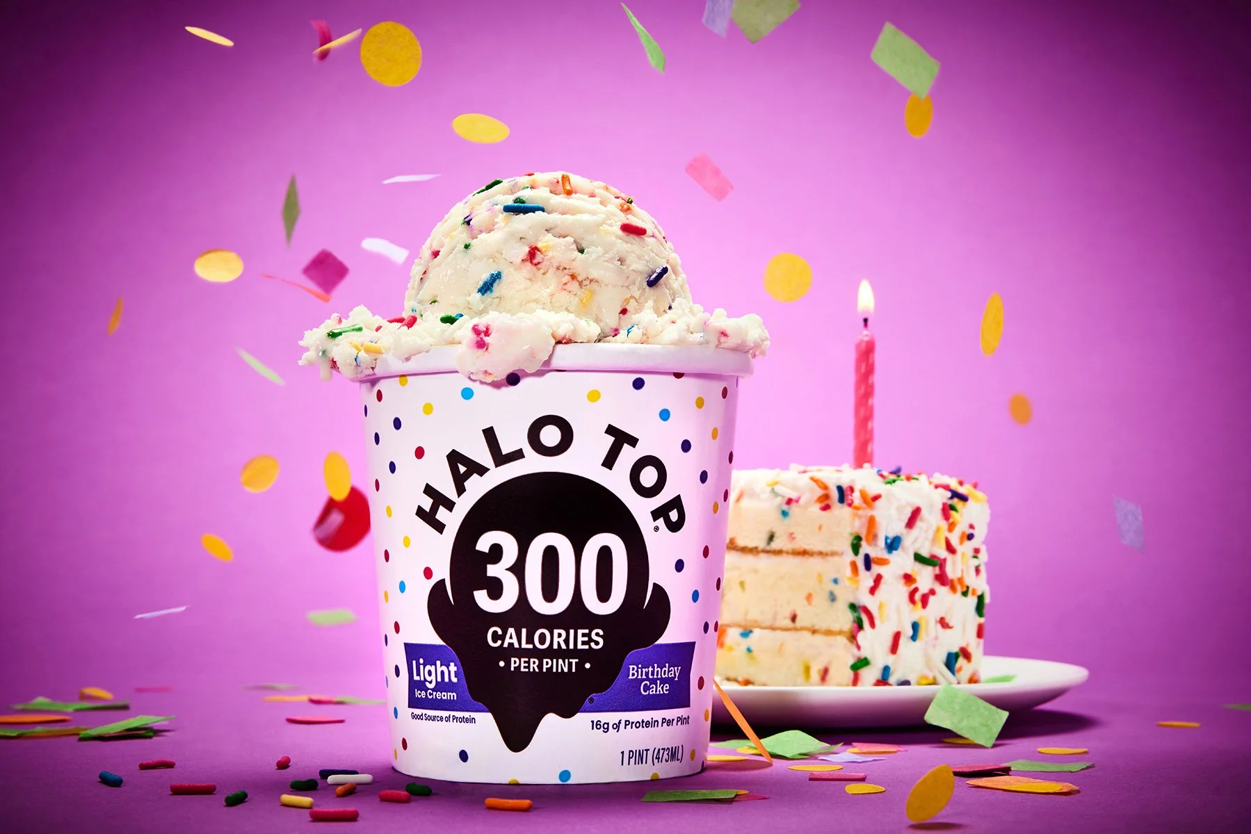 A pint of birthday cake flavored ice cream with rainbow sprinkles and colorful confetti falling around, set against a purple background. There is also a slice of birthday cake with rainbow sprinkles on a plate nearby, and a lit birthday candle next t