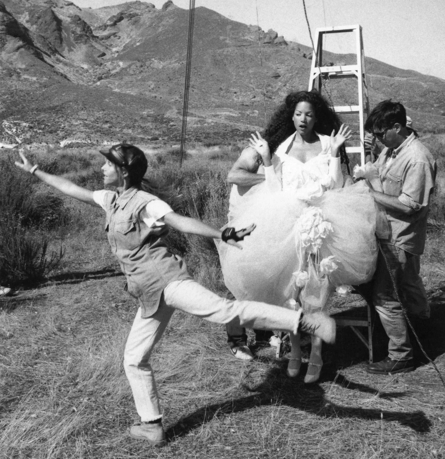 A group of young people outdoors near a mountainous area, with one girl dressed in a large, fluffy dress, and others assisting or posing with her, possibly during a photoshoot.
