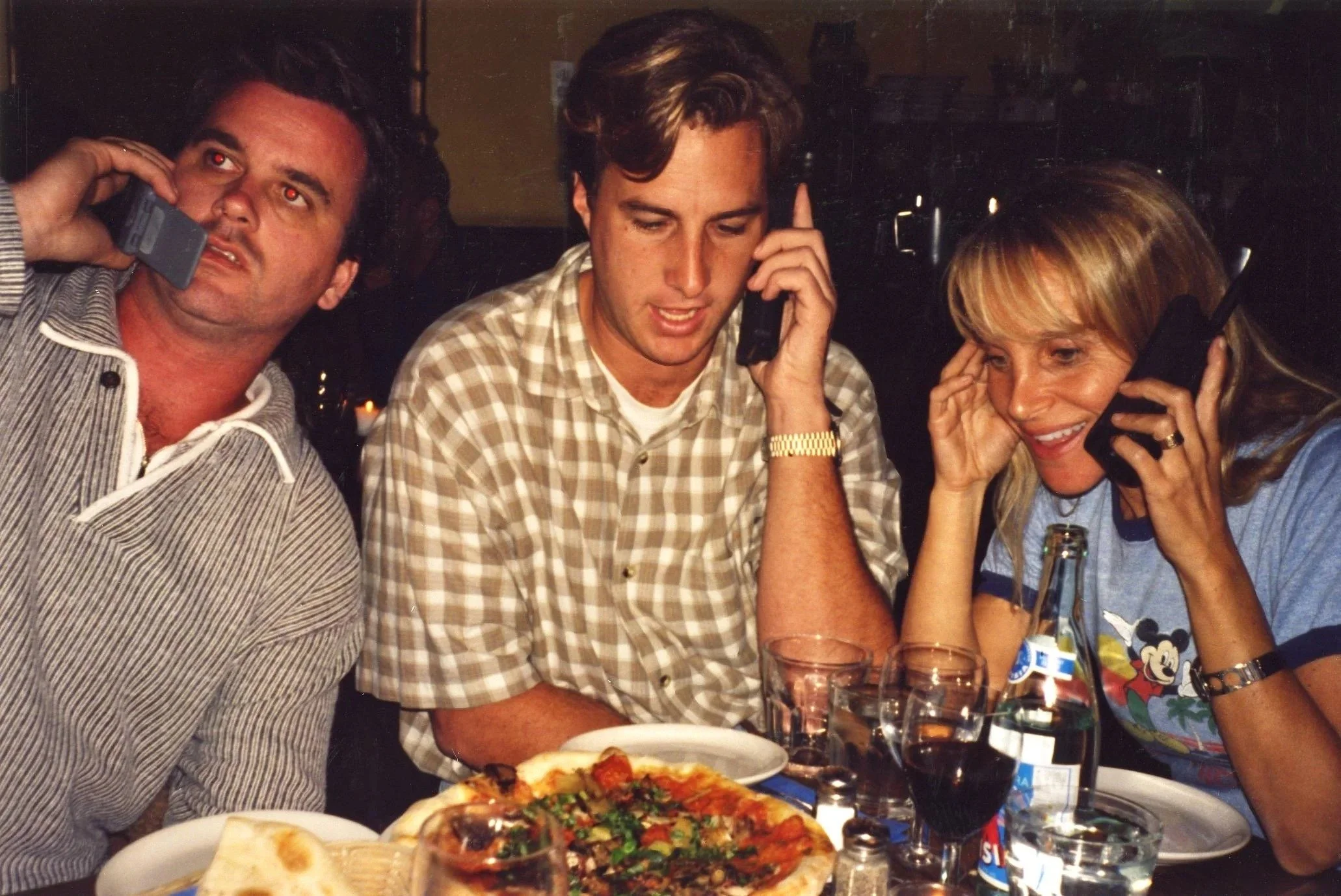 Three people, two men and one woman, are sitting at a table in a restaurant, each on a cell phone. The woman is smiling and wearing headphones. The man in the middle is wearing a checkered shirt, also on the phone, while the man on the left is holdin