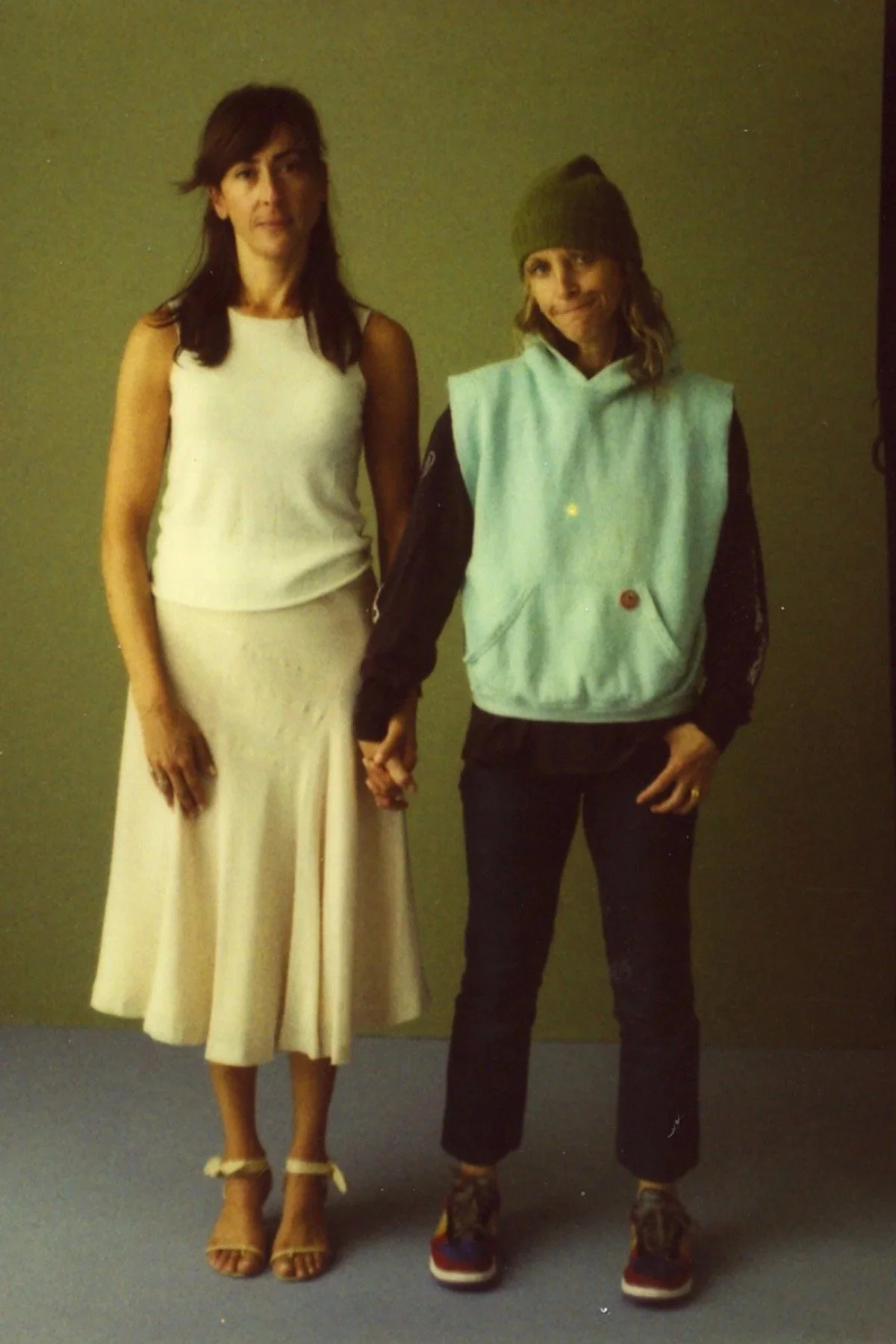 A woman in a white sleeveless top and beige skirt holding hands with a woman in a green beanie, black jacket, and dark pants against a plain background.