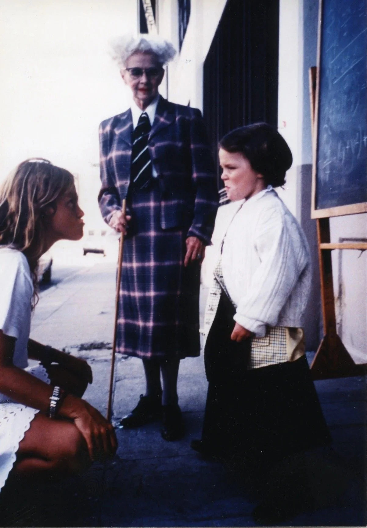An older woman with white hair, wearing sunglasses and a plaid dress, standing near two young girls who are squinting and looking at her skeptically, outdoors in front of a chalkboard.