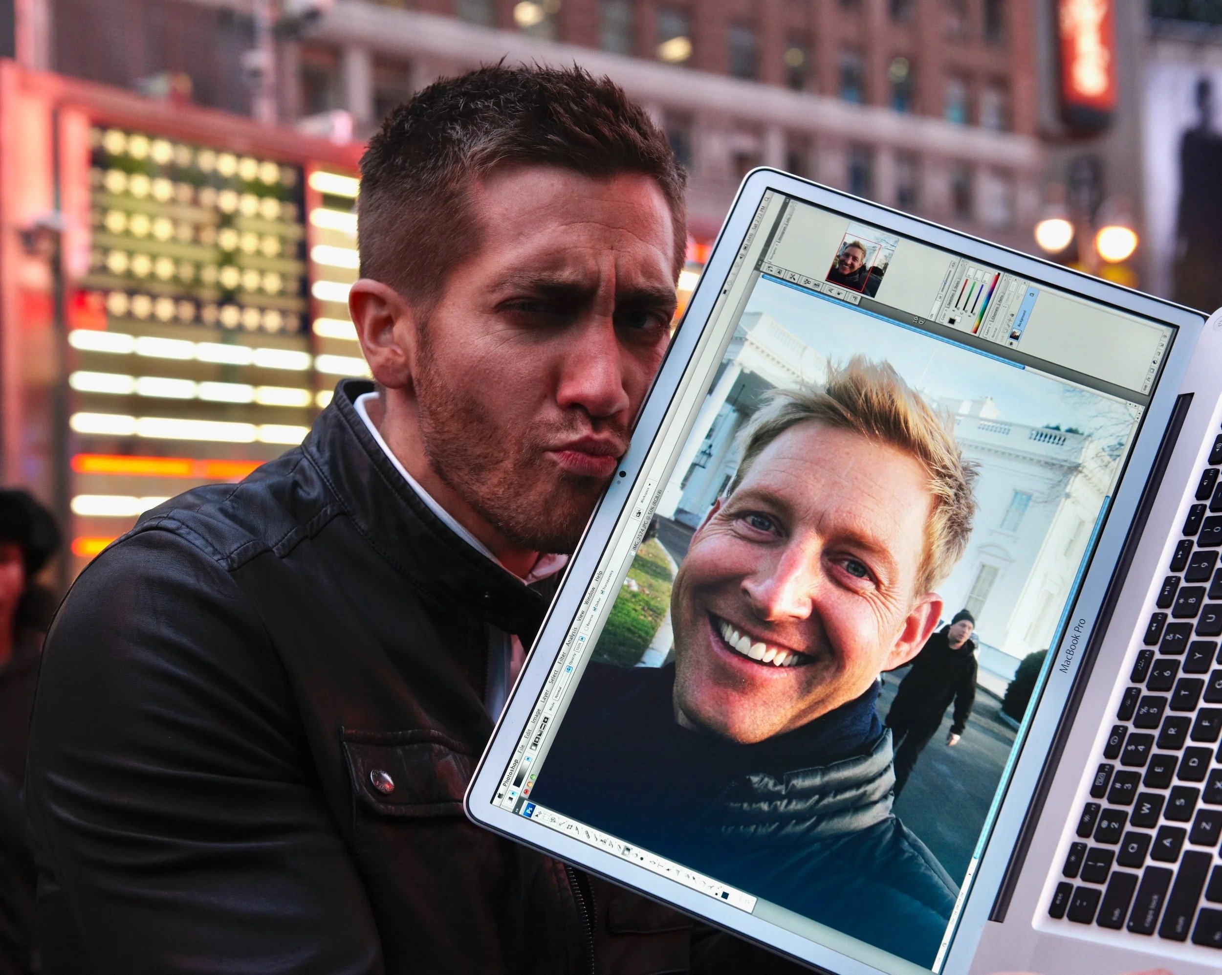 A man with short dark hair holding a laptop displaying a smiling man's face on the screen, with urban buildings in the background.