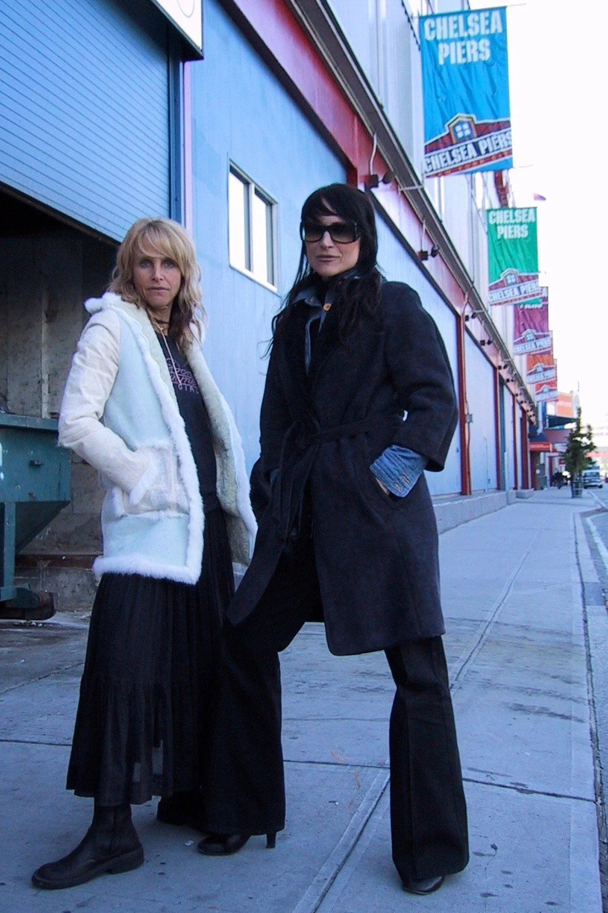Two women standing on a sidewalk outside a building with colorful banners that read 'Chelsea Piers.' One woman has blonde hair, is wearing a white fur-lined coat, and a long black skirt. The other woman has dark hair, is wearing glasses, and a black 
