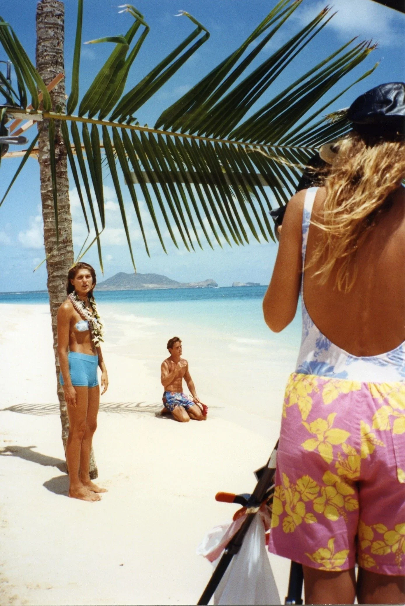 People on a sandy beach with palm trees, ocean, and distant islands in the background. One woman is standing in the foreground, another woman with long hair in the middle, and a man kneeling on the sand, all in swimwear.