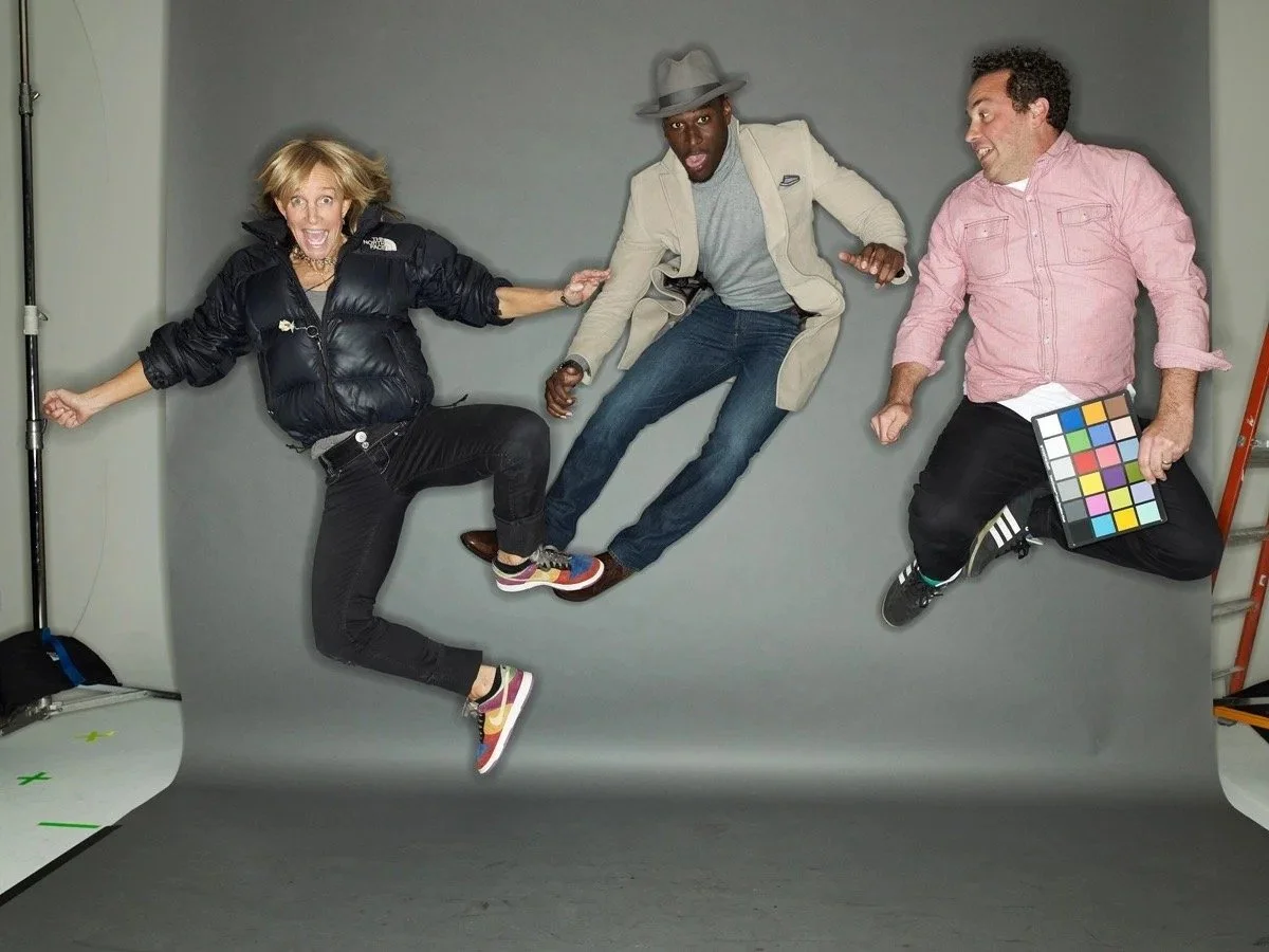 Three people appear to be floating or jumping in mid-air against a gray background, with one holding a color calibration chart.