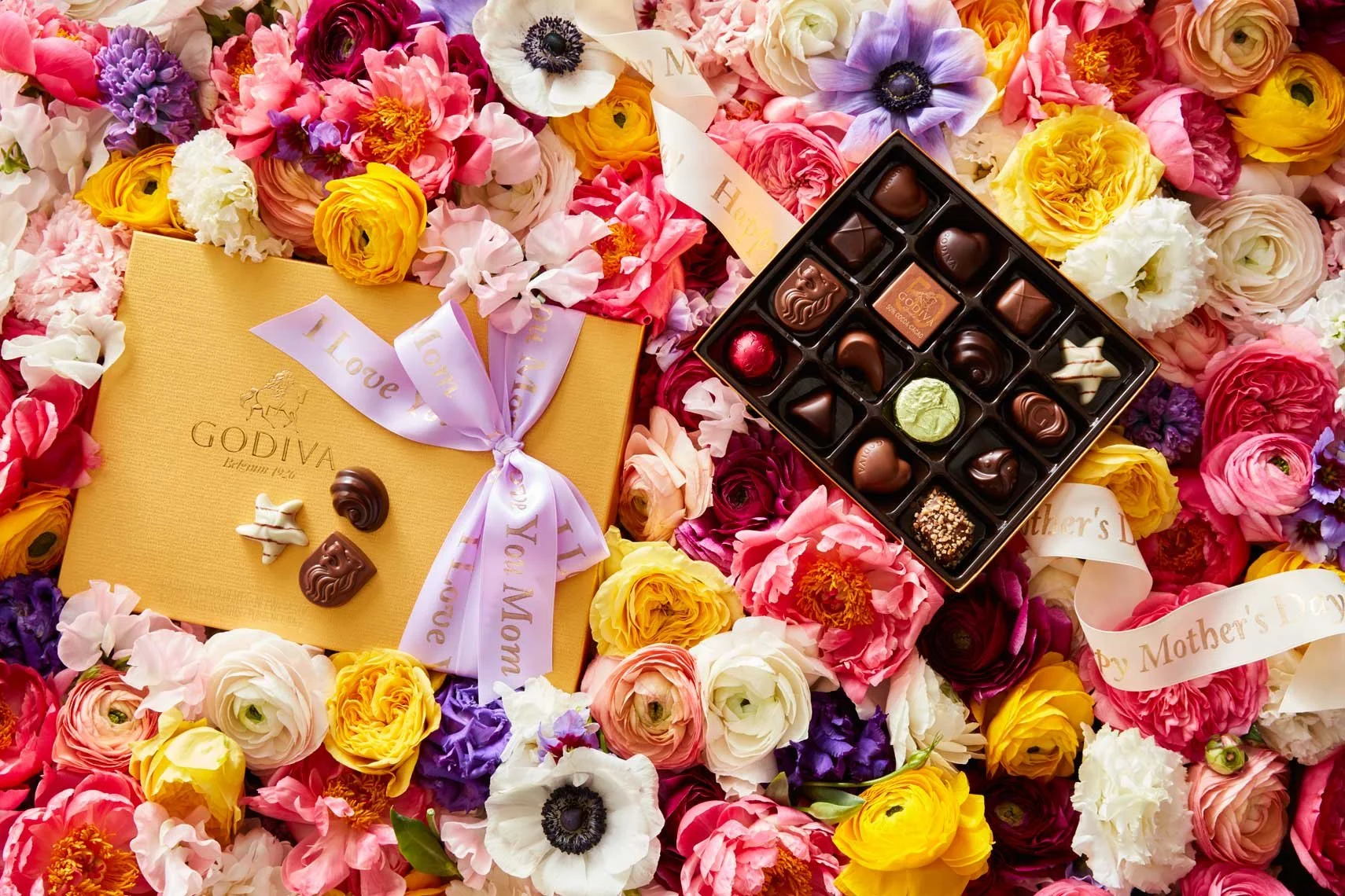 A colorful napkin of pink and purple roses surrounds a box of assorted chocolates from Godiva, with some chocolates placed on a yellow gift box with a pink ribbon and Mother's Day and love messages on the ribbons.