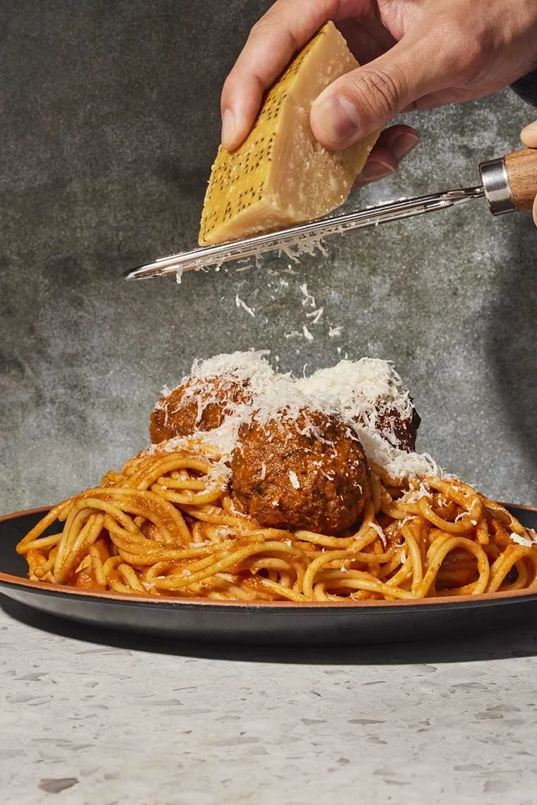 Person grating cheese over a plate of spaghetti and meatballs.