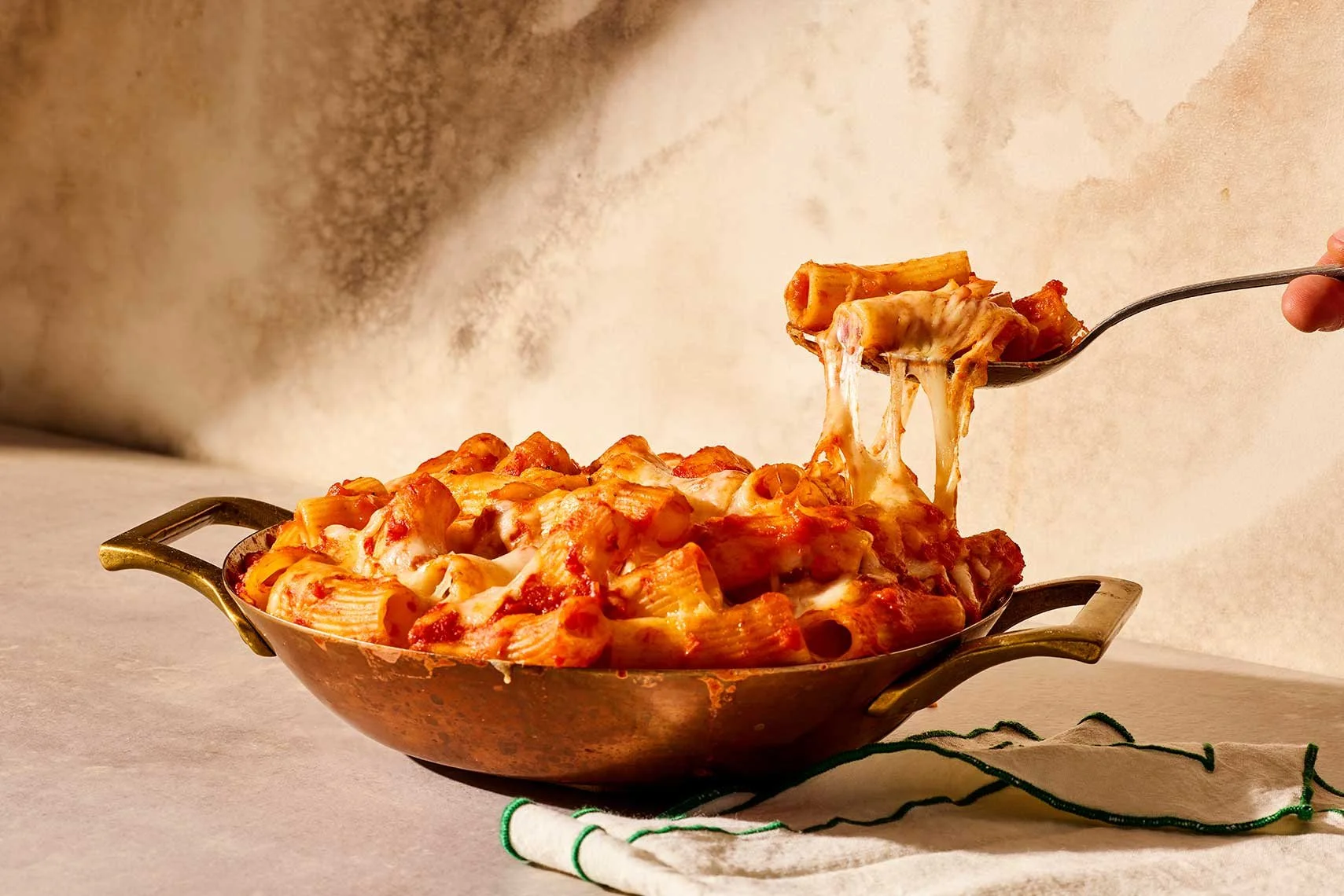 A cast iron skillet filled with cooked pasta, covered in tomato sauce and melted cheese, with a person lifting a portion of cheesy pasta on a fork.