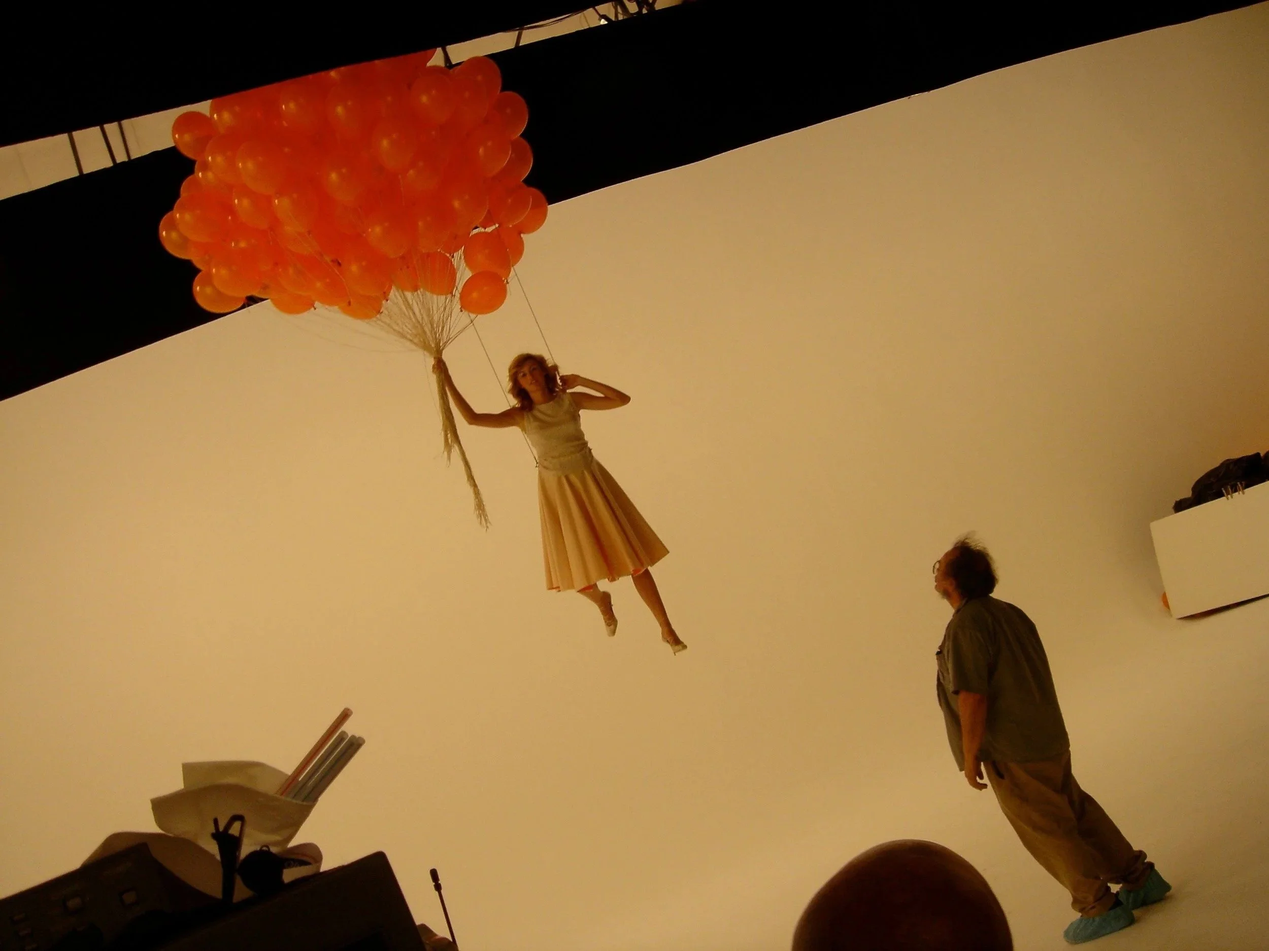A woman holding and floating on a cluster of orange balloons in an indoor space with warm beige walls and black ceiling beams.