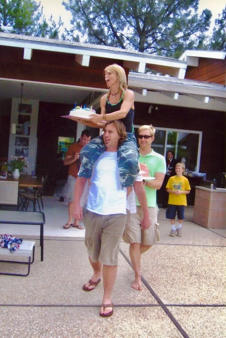 People celebrating at an outdoor birthday party, with a woman holding a birthday cake with candles on top, seated on a man's shoulders.