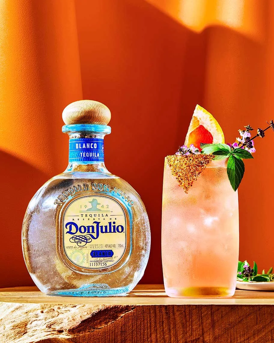 A bottle of Don Julio Blanco tequila and a cocktail garnished with a slice of orange, herbs, and flowers on a wooden surface against an orange background.